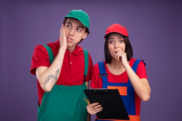 Pareja joven ansiosa en uniforme de trabajador de la construcción y gorra chica sosteniendo lápiz y portapapeles mordiendo el dedo mirando a la cámara chico manteniendo la mano en la cara mirando al lado aislado en la pared púrpura