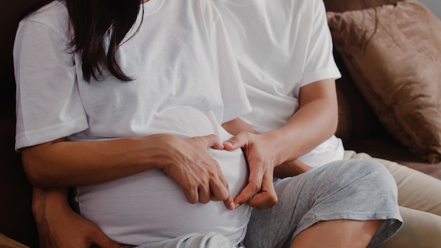 Pareja joven asiática embarazada haciendo muestra del corazón sosteniendo el vientre. Mamá y papá se sienten felices sonriendo pacíficos mientras cuidan bebé, embarazo acostado en el sofá en la sala de estar en casa.