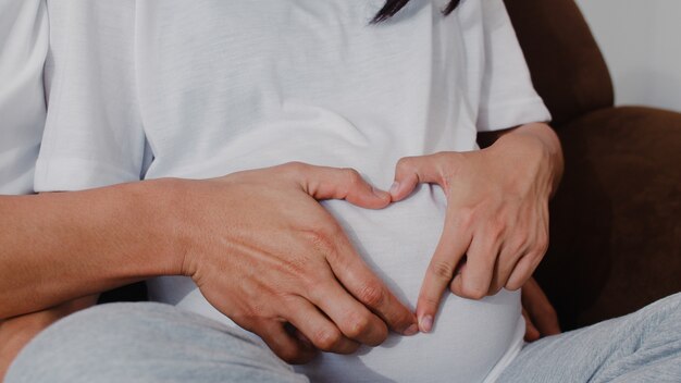 Pareja joven asiática embarazada haciendo muestra del corazón sosteniendo el vientre. Mamá y papá se sienten felices sonriendo pacíficos mientras cuidan bebé, embarazo acostado en el sofá en la sala de estar en casa.