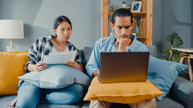 Pareja joven asiática hombre y mujer se sientan en el sofá con documentos de verificación de portátil