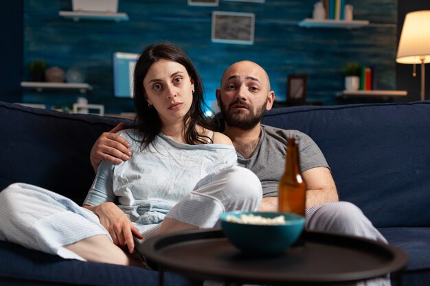 Pareja joven asombrada confundida viendo un documental con expresión facial conmocionada, comiendo palomitas de maíz sentado en el sofá. Adultos concentrados mirando televisión a altas horas de la noche disfrutando de tiempo libre