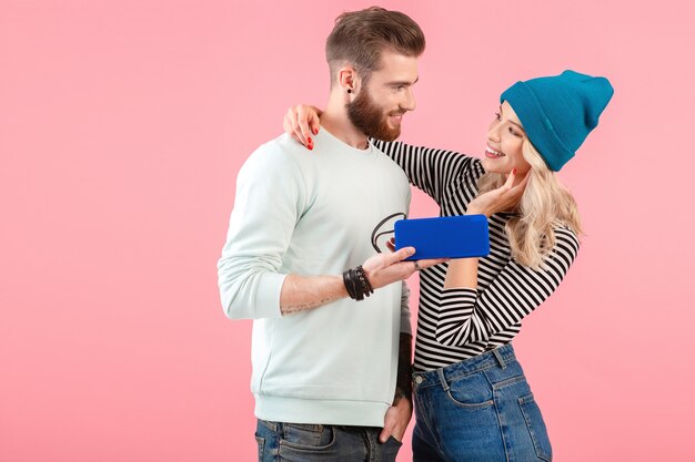 Pareja joven y atractiva escuchando música en un altavoz inalámbrico con un traje elegante y fresco sonriendo feliz estado de ánimo positivo posando sobre fondo rosa