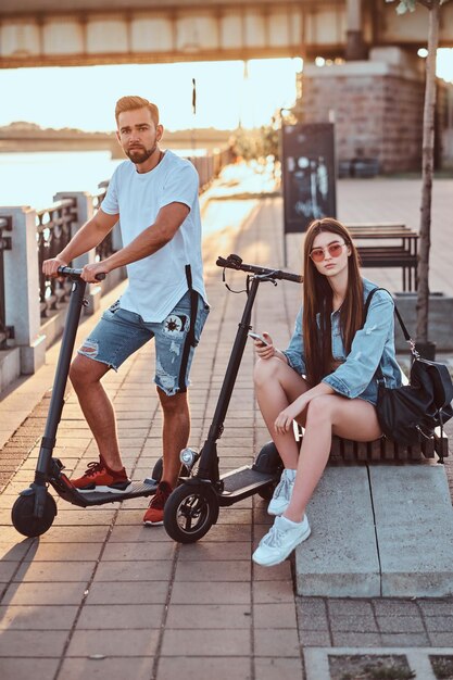 Una pareja joven y atractiva se relaja a orillas del río con sus scooters eléctricos. Hay un día soleado y brillante.