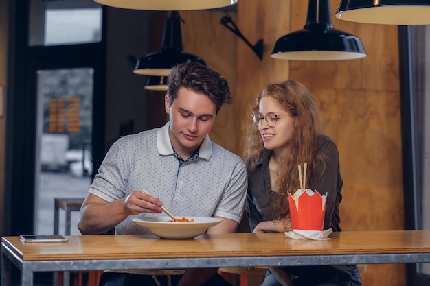 Una pareja joven y atractiva con ropa informal comiendo fideos picantes en un restaurante asiático.