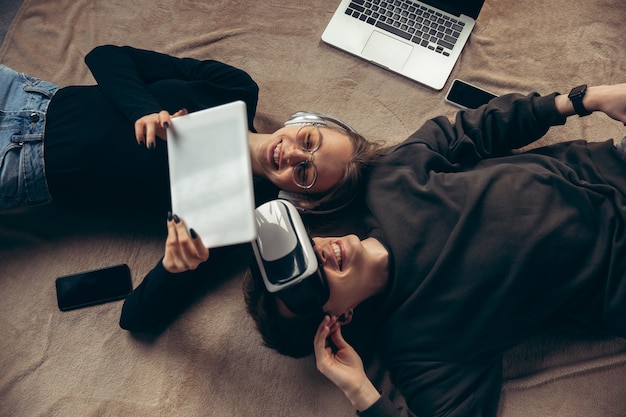 Pareja joven atractiva usando dispositivos juntos, tableta, computadora portátil, teléfono inteligente, auriculares inalámbricos. Comunicación, concepto de gadgets. Tecnologías que conectan a las personas en el autoaislamiento. Estilo de vida en casa.