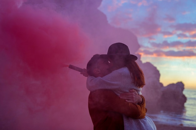 Pareja joven con bomba de humo besándose en la orilla del mar