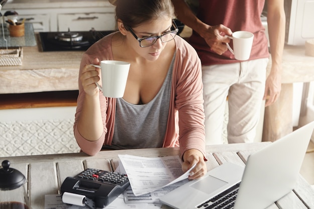 Pareja joven calculando los gastos familiares en casa. Mujer con gafas pagando facturas de servicios públicos en línea, tomando café o té, sentada en la cocina con documentos y calculadora, mirando la pantalla del portátil