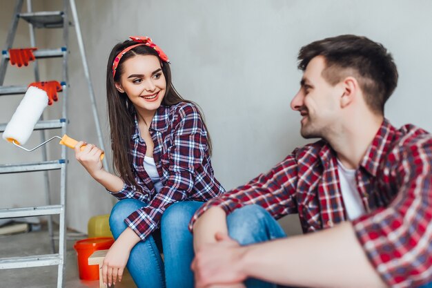 Pareja joven cansada está sentada en el suelo con un pincel y un cubo mientras hace reparaciones en casa