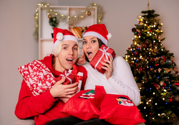 Pareja joven en casa en Navidad con gorro de Papá Noel sentado en un sillón sosteniendo sacos y paquetes de regalo de Navidad impresionó a chico y chica sorprendida, ambos mirando a cámara en la sala de estar