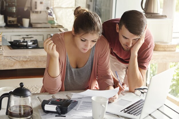 Pareja joven casada con muchas deudas haciendo papeleo juntos, revisando sus facturas, planificando el presupuesto familiar y calculando las finanzas en la mesa de la cocina con papeles