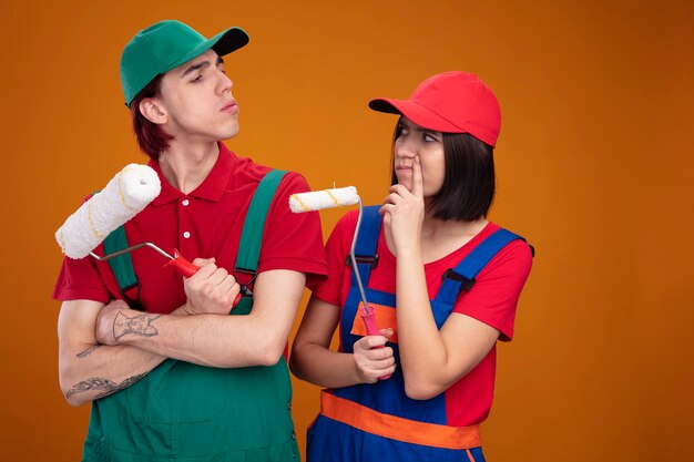 Pareja joven con el ceño fruncido en uniforme de trabajador de la construcción y gorra mirando el uno al otro sosteniendo el rodillo de pintura chico de pie con la chica de postura cerrada tocando la cara con el dedo aislado en la pared naranja