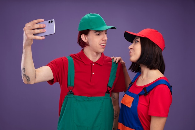 Pareja joven chico alegre chica complacida en uniforme de trabajador de la construcción y gorra tomando selfie juntos mirando el uno al otro chica manteniendo la mano sobre el hombro del chico aislado en la pared púrpura