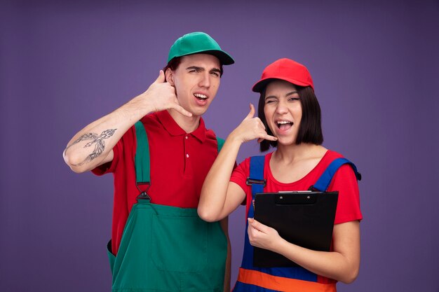 Pareja joven chico seguro y chica alegre en uniforme de trabajador de la construcción y gorra haciendo gesto de llamada chica sosteniendo lápiz y guiño de portapapeles