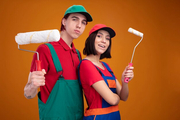 Pareja joven chico seguro y chica emocionada en uniforme de trabajador de la construcción y gorra de pie en vista de perfil sosteniendo rodillo de pintura mirando a cámara aislada en pared naranja