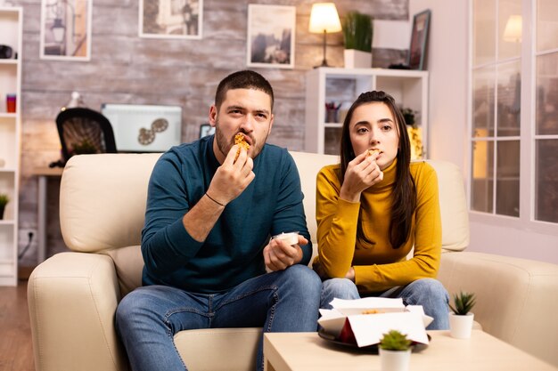 Pareja joven comiendo pollo frito frente al televisor en la sala de estar