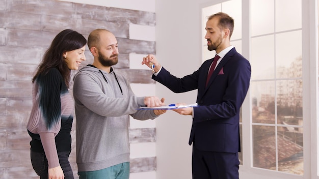 Pareja joven convirtiéndose en propietarios después de firmar documentos con un agente inmobiliario en traje de negocios. Agente dando llaves a pareja.