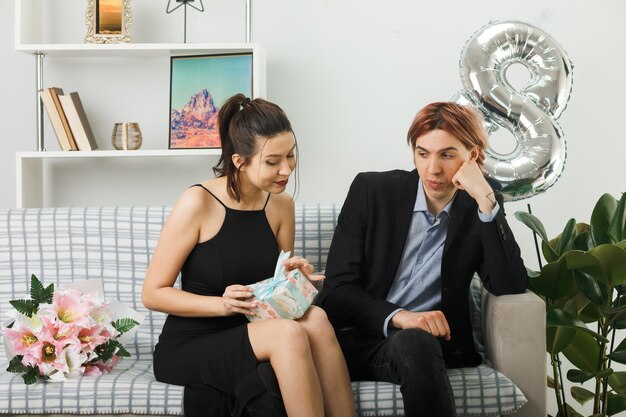 Pareja joven en el día de la mujer feliz sosteniendo y mirando al presente sentado en el sofá en la sala de estar