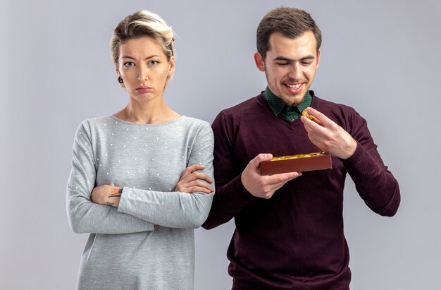 Pareja joven en el día de San Valentín chica descontenta de pie junto al chico con caja de caramelos aislado sobre fondo blanco.