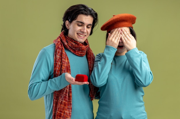 Pareja joven en el día de San Valentín chico con bufanda chica con sombrero chico sonriente dando anillo de bodas a chica aislada sobre fondo verde oliva