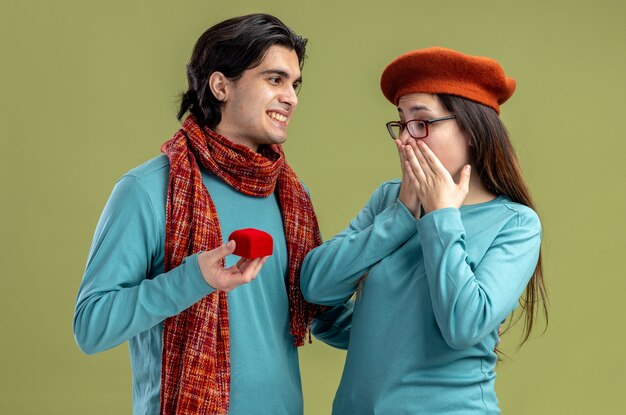 Pareja joven en el día de San Valentín chico con bufanda chica con sombrero chico sonriente dando anillo de bodas a chica sorprendida aislada sobre fondo verde oliva