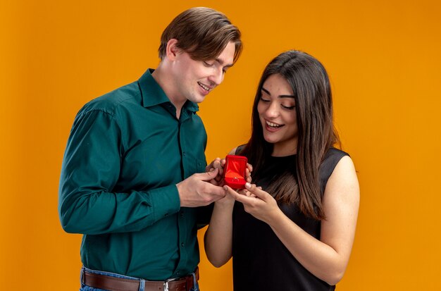 Pareja joven en el día de San Valentín chico complacido dando anillo de bodas a niña sonriente aislada sobre fondo naranja