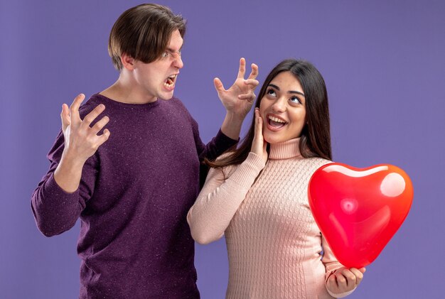 Pareja joven en el día de San Valentín chico enojado mirando a chica emocionada con globo de corazón poniendo la mano en la mejilla aislado sobre fondo azul.