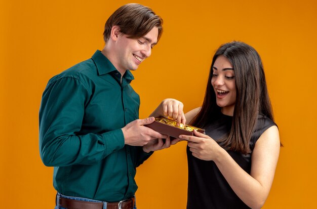 Pareja joven en el día de San Valentín chico sonriente dando caja de caramelos a niña sorprendida aislada sobre fondo naranja