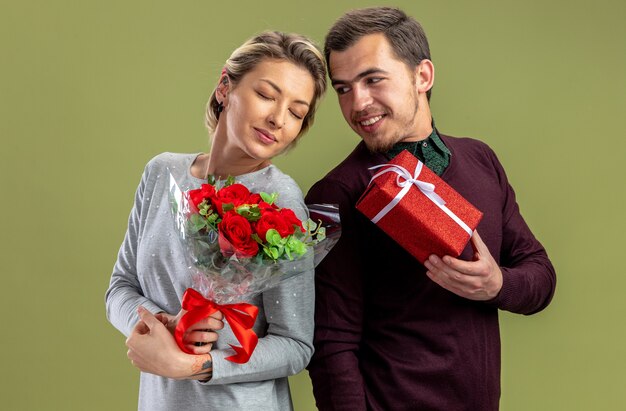 Pareja joven en el día de San Valentín chico sonriente dando caja de regalo a niña complacida con ramo aislado sobre fondo verde oliva