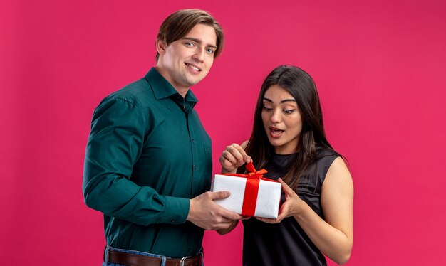 Pareja joven en el día de San Valentín chico sonriente dando caja de regalo a niña sorprendida aislada sobre fondo rosa