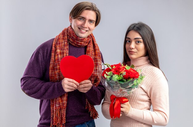 Pareja joven en el día de San Valentín chico sonriente sosteniendo una caja en forma de corazón chica complacida sosteniendo un ramo aislado sobre fondo blanco.