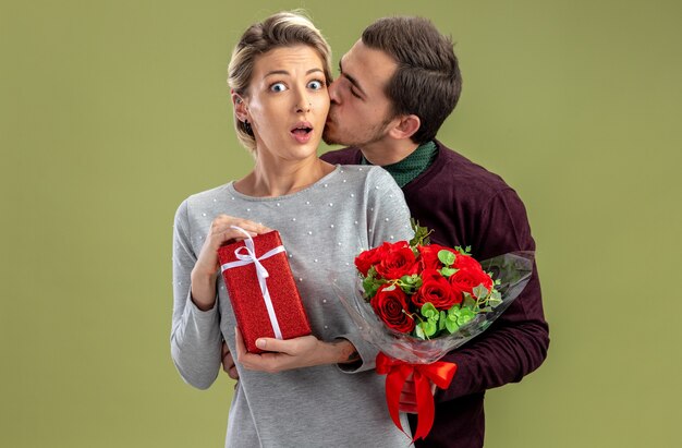 Pareja joven en el día de San Valentín chico sosteniendo ramo besando a chica sorprendida con caja de regalo aislada sobre fondo verde oliva