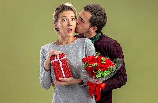 Pareja joven en el día de San Valentín chico sosteniendo ramo besando a chica sorprendida con caja de regalo aislada sobre fondo verde oliva