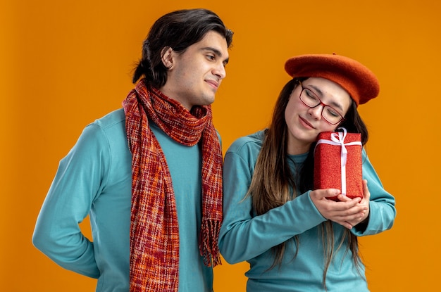 Pareja joven en el día de San Valentín feliz chico con bufanda mirando a la chica con caja de regalo aislada sobre fondo naranja