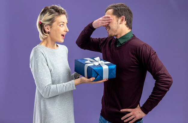 Pareja joven en el día de San Valentín niña sonriente dando caja de regalo a chico aislado sobre fondo azul.