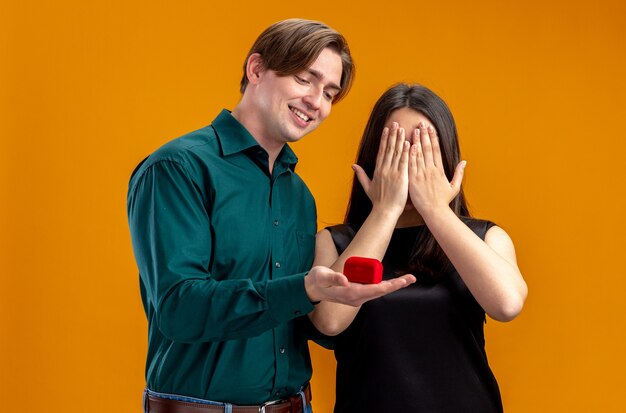 Pareja joven en el día de San Valentín smilin guy dando anillo de bodas a niña aislada sobre fondo naranja