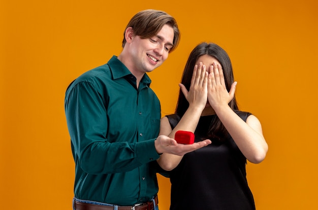 Pareja joven en el día de San Valentín smilin guy dando anillo de bodas a niña aislada sobre fondo naranja