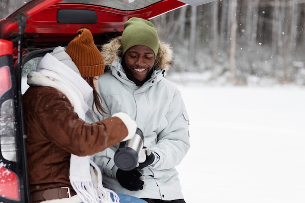 Pareja joven disfrutando de bebidas calientes durante un viaje por carretera en invierno en el maletero del coche