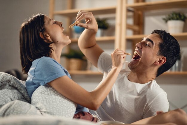 Pareja joven divirtiéndose mientras se alimentan mutuamente con galletas en el dormitorio