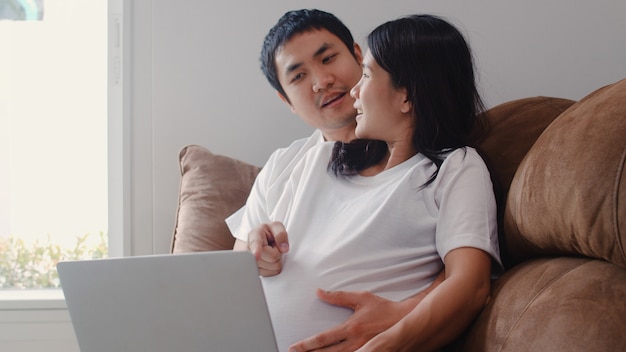 Pareja joven embarazada de Asia utilizando información de embarazo de búsqueda portátil. Mamá y papá se sienten felices sonriendo positivos y tranquilos mientras cuidan a su hijo acostado en el sofá en la sala de estar en casa