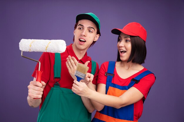 Pareja joven emocionada en uniforme de trabajador de la construcción y chico de gorra sosteniendo y señalando a la chica del rodillo de pintura estirando el pincel mirando el rodillo de pintura aislado en la pared púrpura