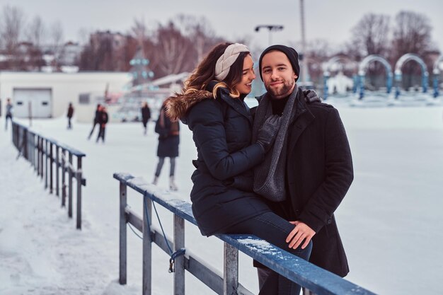 Pareja joven enamorada, cita en la pista de hielo, una chica sentada en una barandilla y abrazando a su novio.