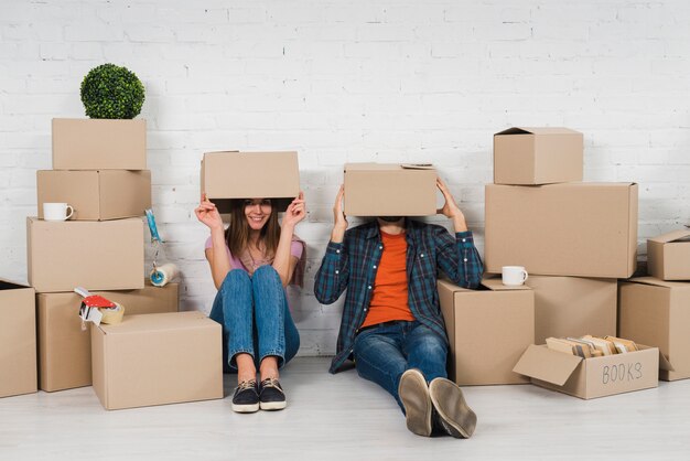 Pareja joven escondiendo su cara sentada entre las cajas de cartón en su nueva casa