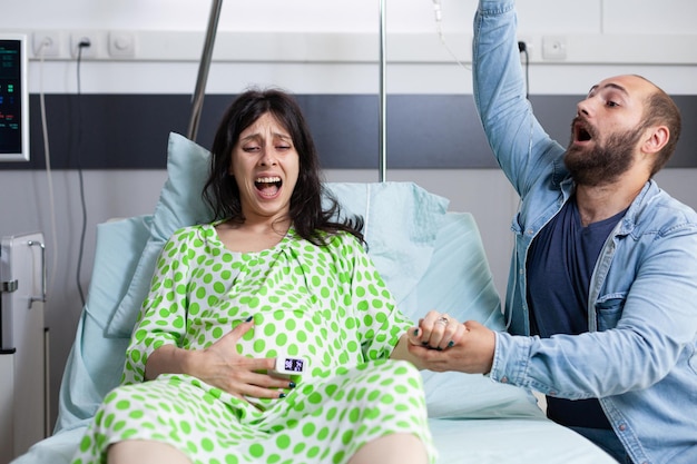 Foto gratuita pareja joven esperando un bebé en la cama de un hospital en un centro de salud. una mujer embarazada tiene contracciones dolorosas y se pone de parto mientras su marido entra en pánico por el parto.