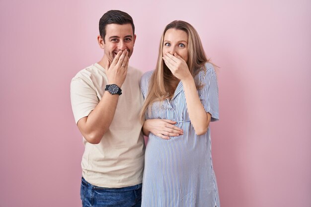 Una pareja joven esperando un bebé parado sobre un fondo rosado riéndose y riéndose avergonzada cubriendo la boca con chismes de manos y concepto de escándalo