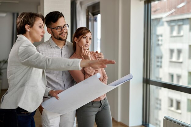 Pareja joven examinando planos con un agente inmobiliario mientras compra una casa nueva