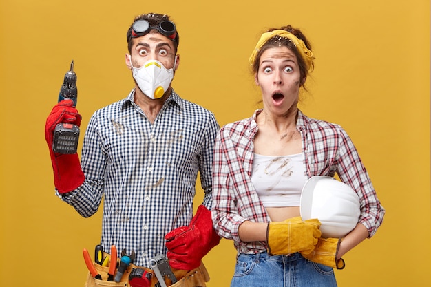 Pareja joven de la familia vistiendo ropa protectora mientras hacen reparaciones en su apartamento sosteniendo un taladro y un casco tener una mirada de sorpresa tener miedo de hacer mucho trabajo y tener la cara sucia