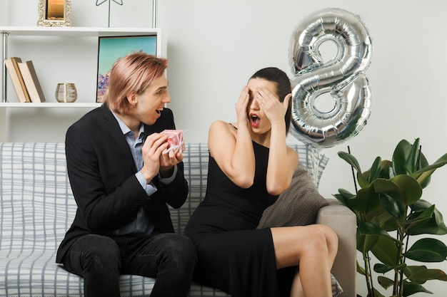 Pareja joven en feliz día de la mujer chico emocionado da presente a chica sorprendida cubrió los ojos con las manos sentadas en el sofá en la sala de estar