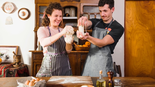 Pareja joven feliz jugando con masa de pie detrás de la mesa de madera