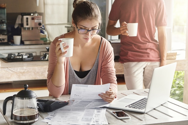 Pareja joven frente a problemas financieros, gestión del presupuesto familiar en la cocina. Mujer casual en vasos tomando café y sosteniendo el trozo de papel