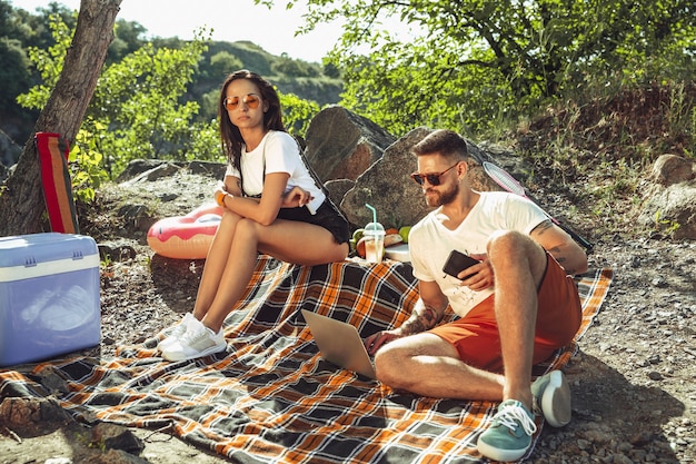 Pareja joven haciendo un picnic en la orilla del río en un día soleado. mujer y hombre pasando tiempo juntos en la naturaleza. divertirse, comer, jugar y reír. concepto de relación, amor, verano, fin de semana.